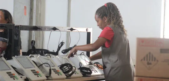 colaboradora da Justiça Eleitoral manuseia urna eletrônica disposta em uma fila com outros dispositivos de votação.